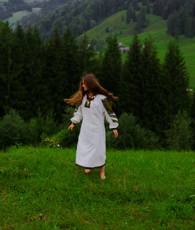 girl with loose hair in an embroidery with a bouquet of wild flowers sits on green grass against the background of the carpathian mountainsの写真素材