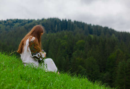 girl with loose hair in an embroidery with a bouquet of wild flowers sits on green grass against the background of the carpathian mountainsの写真素材