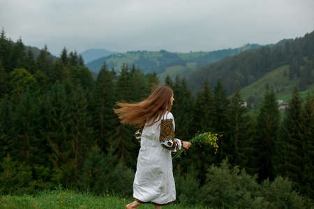 girl with loose hair in an embroidery with a bouquet of wildflowers happily runs along the green in the carpathian mountainsの写真素材