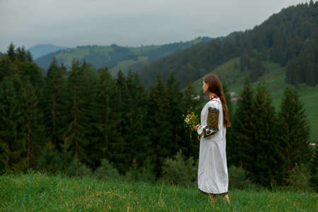 girl in embroidery with a bouquet of wild flowers stands on green grass in the carpathian mountainsの写真素材