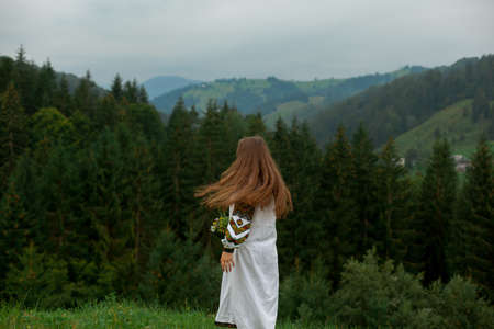 girl with loose hair in an embroidery with a bouquet of wildflowers happily runs along the green in the carpathian mountainsの写真素材
