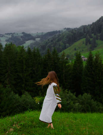 girl with loose hair in an embroidery with a bouquet of wildflowers happily runs along the green in the carpathian mountainsの写真素材