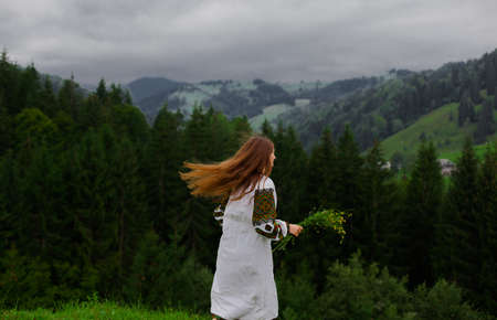 girl with loose hair in an embroidery with a bouquet of wildflowers happily runs along the green in the carpathian mountainsの写真素材