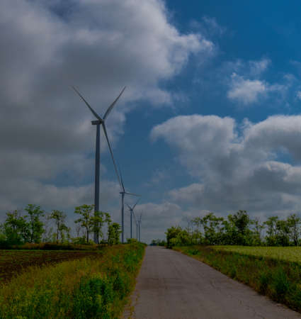 Under the blue sky with white clouds, the propeller of the green electricity generator is mounted on the side of the road in the fieldの写真素材