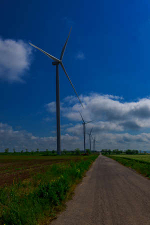 Under the blue sky with white clouds, the propeller of the green electricity generator is mounted on the side of the road in the fieldの写真素材