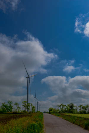 Under the blue sky with white clouds, the propeller of the green electricity generator is mounted on the side of the road in the fieldの写真素材