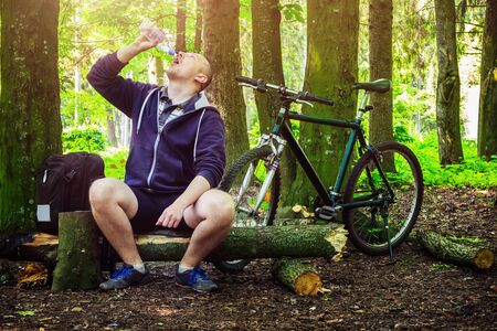 Cyclist with backpack, young man drinking water from bottle, in beautiful forest, summertime journeyの写真素材