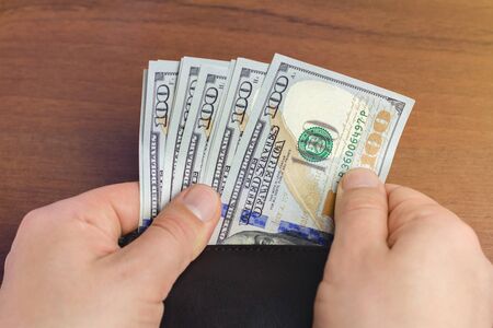 Close-up of male hand taking hundred dollar banknotes from wallet, wooden background, money and...