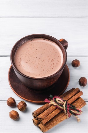 Hot cocoa with milk in brown clay cup, hazelnut and cinnamon sticks on table of white wooden planks.の写真素材