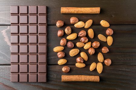 Bitter, dark chocolate bar, almond, hazelnut and cinnamon stick on wooden background, top view.の写真素材