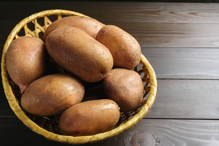 Uncooked, fresh crop of potatoes in a wicker basket on wooden table of dark brown planks background.の写真素材