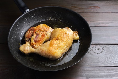 Two roasted chicken legs in black frying pan on dark wooden planks close-up view.の写真素材