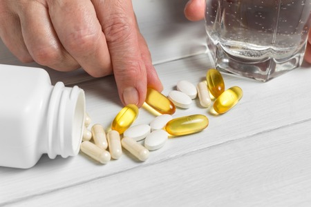 Woman hands takes yellow capsules of omega 3, white pills of calcium, glucosamine supplements, glass of water and plastic bottle on wooden planks table.の写真素材