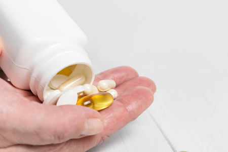 Woman hands hold vitamin complex and dietary supplement, pours from white bottle into palm yellow capsules of omega 3, white pills of calcium and glucosamine on wooden planks table.の写真素材