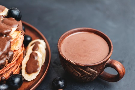 Hot chocolate drink in a cup and pancakes with banana, chocolate sauce and grapes in plate, on dark background.の写真素材