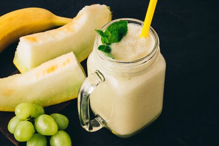 Melon smoothie in a jar with fresh mint leaves, banana and grape on dark background.の写真素材