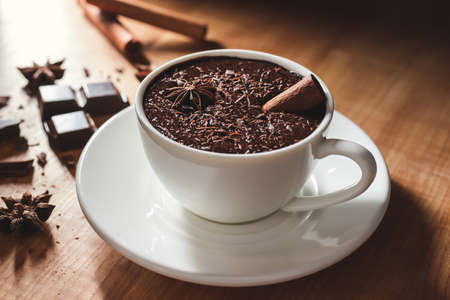 Hot chocolate drink in white cup, broken chocolate, cinnamon stick and star anise on a wooden table.の写真素材