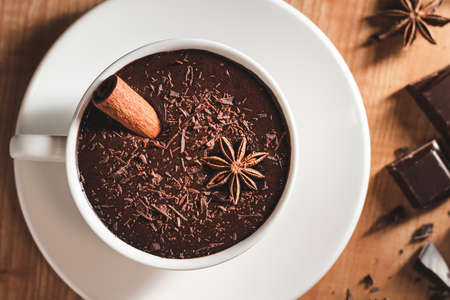 Hot chocolate drink in white cup, broken chocolate, cinnamon stick and star anise on a wooden table,...
