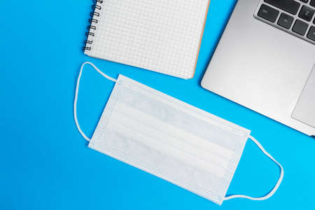 Flat lay composition with laptop, medical face mask, notepad on blue background top view with copy space. Concept of safety work in coronavirus pandemic.の写真素材