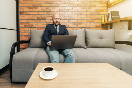 Bearded bald man, businessman or freelancer sitting on sofa and working on laptop from home, modern interior loft design.の写真素材