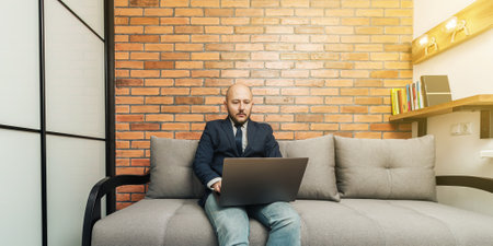 Bearded bald man, businessman or freelancer sitting on sofa and working on laptop from home, modern interior loft design.の写真素材