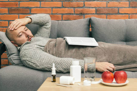 Sick man with book lies on sofa and has symptoms of infection coronavirus, covid-19 or flu illness, table with medications and pills in the room.の写真素材