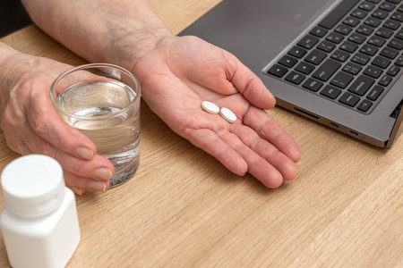 White pills of painkiller or antibiotic for treatment on senior woman hand palm, glass with water, medicines and vitamin supplements concept, close-up view.の写真素材
