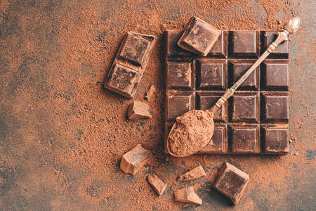 Brown cocoa powder in the spoon on chocolate bar and chopped chocolate cubes, top view on dark...