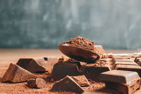 Brown cocoa powder in the spoon and chopped chocolate cubes, on dark background with copy space.の写真素材