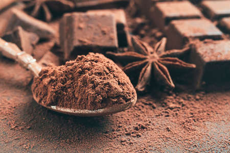 Brown cocoa powder in the spoon, chopped chocolate cubes and star anise on dark background.の写真素材