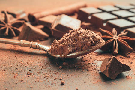 Brown cocoa powder in the spoon, chopped chocolate cubes and star anise on dark background.