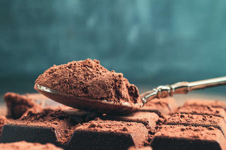 Brown cocoa powder in the spoon on chocolate bar, chopped chocolate cubes on dark background with...