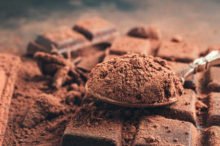 Brown cocoa powder in the spoon on chocolate bar, chopped chocolate cubes on dark background.