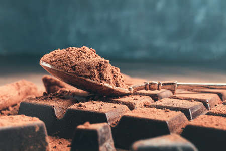 Brown cocoa powder in the spoon on chocolate bar, chopped chocolate cubes on dark background with copy space.の写真素材
