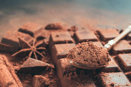 Brown cocoa powder in the spoon on chocolate bar, chopped chocolate cubes on dark background with...