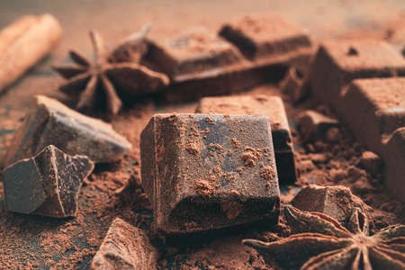 Chopped chocolate cubes with cocoa powder and star anise on dark background, close-up view.