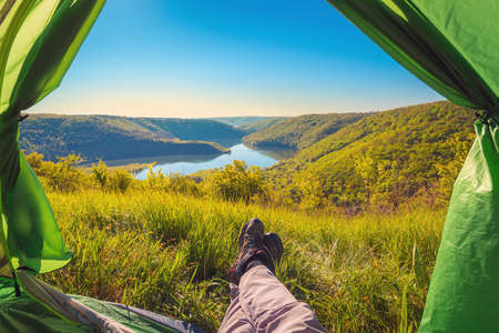 POV view, feet in the trekking shoes of the resting traveler in the camping tent, view to the mountain canyon and river, scenic nature background, adventure concept.の写真素材