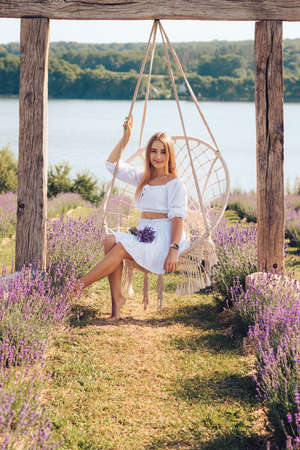 Portrait of young pretty, charming female with lavender bouquet sitting on swing, outdoors at flower field in the summer.の写真素材