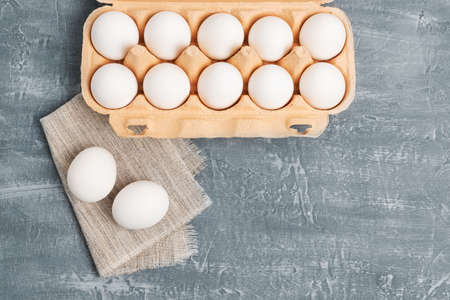 White chicken eggs in carton box on grey textured background, top view.