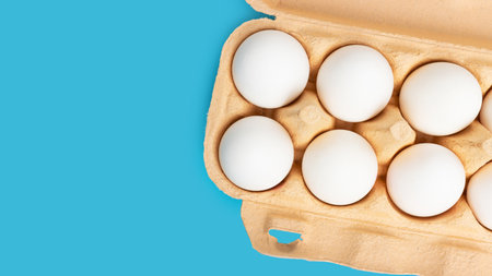 White chicken eggs in carton box on blue background, top view.