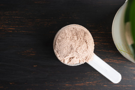 Whey chocolate protein powder in a plastic measuring spoon on a dark wooden background, top view.の写真素材