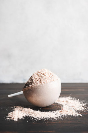 Whey chocolate protein powder in a plastic measuring spoon on a dark wooden board and white...