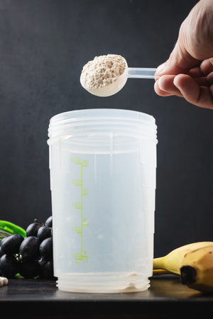 Male hand puts into a shaker scoop portion of whey, soy protein powder. White capsules of amino acids, vitamins, protein chocolate bar and bananas on a dark wooden board, bodybuilding food supplementsの写真素材