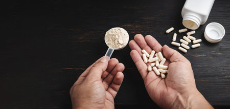 Male hands holds a scoop of whey or soy protein powder and white capsules of amino acids, vitamins, creatine, bodybuilding food supplements, sports nutrition on a dark wooden board.の写真素材