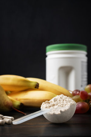 Scoop of whey or soy protein powder, white capsules of amino acids, vitamins, creatine, bodybuilding food supplements and sports nutrition. fruits, banana, pear and grape on a dark wooden board.の写真素材