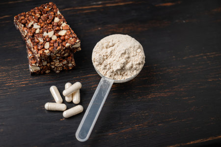 Scoop of whey or soy protein powder, white capsules of amino acids, vitamins, creatine, protein chocolate bar, bodybuilding food supplements, sports nutrition on a dark wooden board.の写真素材