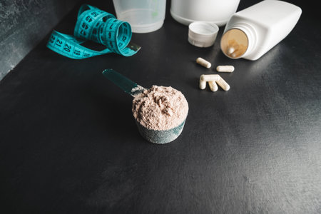 Chocolate whey protein powder in measuring spoon, white capsules of amino acids, vitamins and creatine, measuring tape, plastic shaker on dark background. bodybuilding food supplements.の写真素材