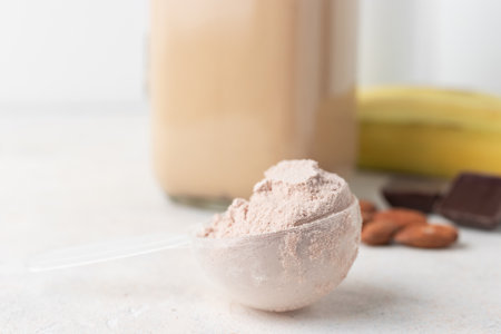 Chocolate whey protein powder in measuring spoon, glass jar of protein milkshake drink or smoothie,...