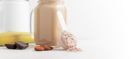 Chocolate whey protein powder in measuring spoon, glass jar of protein milkshake drink or smoothie, bananas, chocolate cubes and almonds on white background. sport nutrition, food supplements.の写真素材