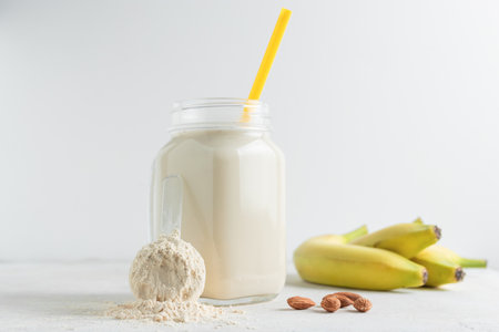 Whey protein powder in measuring spoon, glass jar of protein milkshake drink or smoothie, bananas and almond nuts on a white background. sport nutrition, bodybuilding food supplements.の写真素材
